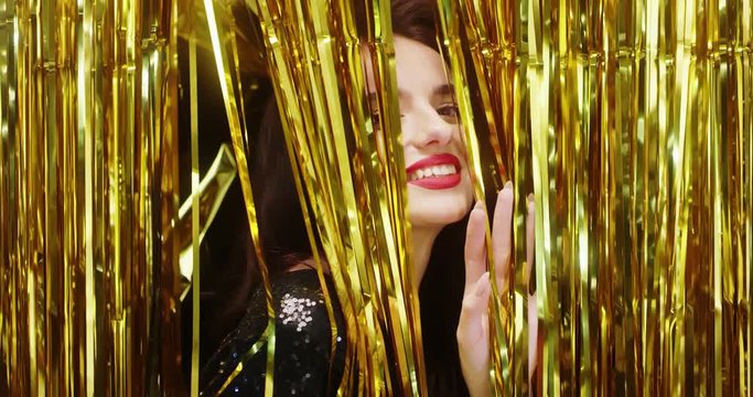 Close up of the attractive Caucasian young lady with red lips peering from the golden shiny confetty strips and smiling joyfully on the New Year Eve celebration. Portrait shot.