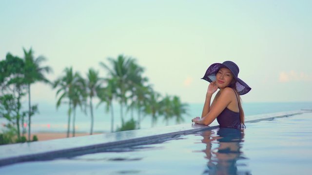 locked medium-wide portrait shot of young adult Asian woman in swimsuit with hat sitting at the edge of infinity pool with defocussed tropical beach and palm trees background copy space