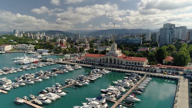 Drone flying up sea port marine station landmark Sochi Russia cityscape skyline resort town modern hotels. Many yachts ships piers parking. Pedestrian embankment. Sunny summer day clouds. Tourism best