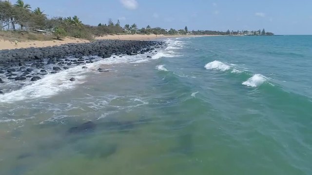 Aerial tracking low over waves breaking on rocky tropical coast, sunny day, slow motion