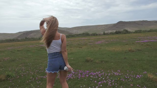 Camera follow hipster young woman in white t-shirt short jeans fluttering long hair. One girl dancing on field of grass portable music speaker smartphone. Happy drunk from life, youth and happiness.