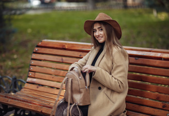 Young attractive woman in autumn clothes sits on bench in city park. Woman dressed in stylish coat...