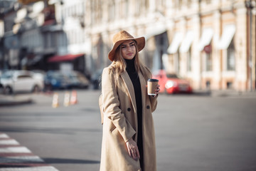 Fototapeta premium Young stylish woman dressed in a coat and felt hat walks around the city and drinks coffee on the go. Autumn time