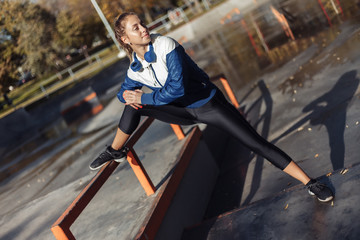 Obraz premium Fit slim woman in sportswear and headphones practicing leg stretching before training in a sports park