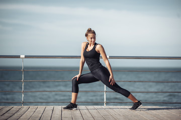 Fototapeta premium Warm up before training. Slim young woman with perfect body doing aerobic exercise on the beach on a bright sunny day.