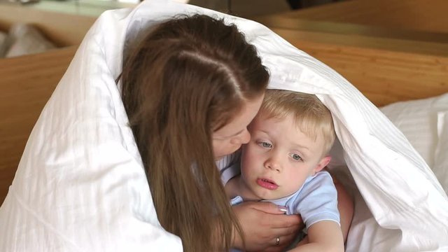 Mother hugs her little sad son on the bed under the blanket. Portrait of a sad baby on the bed, mom hugs and calms the child. Slow motion.