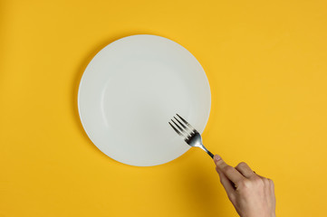 Minimalistic food concept. Female hands holding a fork and an empty white plate on a yellow...