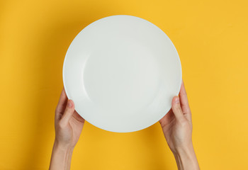 Female hand holds empty white plate on yellow background. Top view.