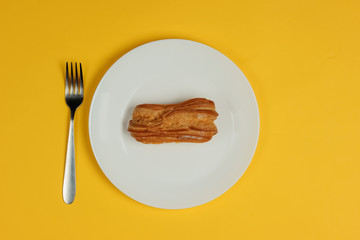 Eclair in plate with fork on yellow background. Top view