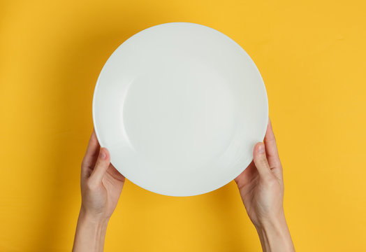 Female Hand Holds Empty White Plate On Yellow Background. Top View.