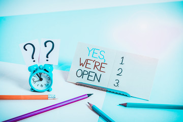 Text sign showing Yes We Re Open. Business photo text answering on client that shop is available at this time Mini size alarm clock beside stationary placed tilted on pastel backdrop