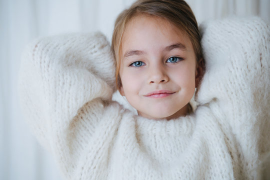 Portrait of a brunette little girl in white fluffy knitted sweater putting her hair in a pony tail. At home, in front of a curtain. She's looking at camera. Close up.