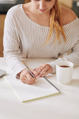 Young woman sitting at kitchen table with cup of coffee and writing affirmations in textbook