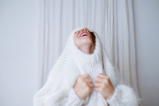 Funny little girl struggling to put on white fluffy knitted sweater. She's squeezing her head out of the collar, pulling fabric with hands. At home, in front of a curtain. Half length.