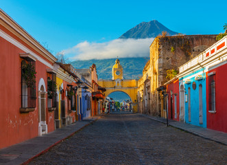 Cityscape of the main street and yellow Santa Catalina arch in the historic city center of Antigua at sunrise with the Agua volcano, Guatemala. - obrazy, fototapety, plakaty