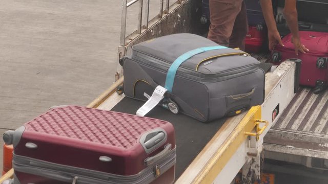 Baggage handlers unload an airplane with conveyor belt delivering suitcases to back of truck for transport to the terminal and waiting passengers. Suitcases moving on belt.