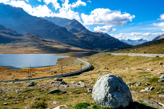 Ferrovia che corre tra le montagne. un grosso masso in primo piano ed un lago sulla sinistra. Cielo blue inteso e pieno di nuvole bianco. Sullo sfondo un ghiacciaio