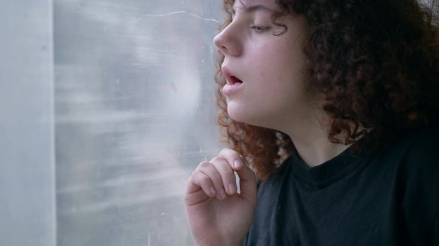 close up portrait of teenage curly brunette girl drawing heart with finger on glass, sad alone, waiting for love, sitting by window indoors, emotions, heartbroken, slow motion