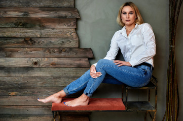 Full-length art portrait of a pretty blonde woman in blue jeans and a white shirt sitting on a...