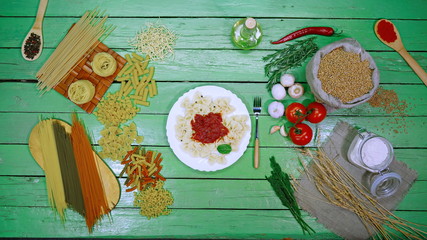 Fototapeta na wymiar Cooked pasta on plate with tomato sauce. Top View. Demonstration of cooked bows pasta and dry pasta. Green table is divided into many types of dry pasta on the left side and vegetables, cherry tomato