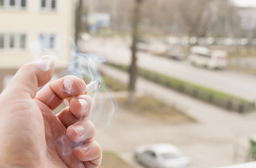 View of the hand of a Smoking man with a Smoking cigarette from the window of the apartment into the yard and the street with passing cars on the road