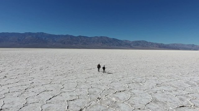 Back view of male and female travelers walking in dry lands of national park in USA carrying rucksacks, friends exploring natural landscape of Badwater basin with white salt on death lake surface