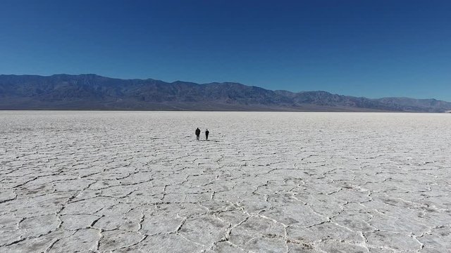 Back view of male and female travelers walking in dry lands of national park in USA carrying rucksacks, friends exploring natural landscape of Badwater basin with white salt on death lake surface