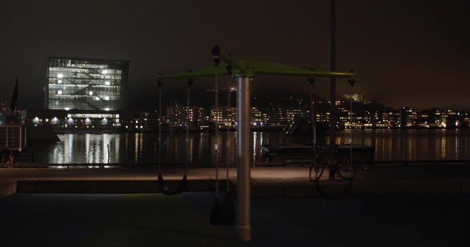 Cinematic wide angle 4K shot with slow parallax motion of empty playground with illuminated Opera Ballet Theatre and Edvard Munch Lambda gallery in background, at night in Bj&oslash;rvika Oslo Norway.