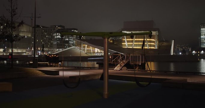 Stabilized wide angle 4K shot with slow parallax motion of empty dark playground and swings with illuminated Opera Theatre and Edvard Munch Lambda museum in background, night in Bj&oslash;rvika Oslo Norway.