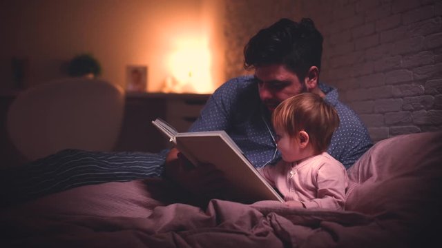 cheerful father is reading the bedtime stories to infant baby girl in bed at night