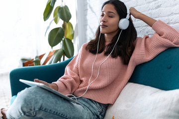 Pretty young woman listening to music with digital tablet and relaxing while sitting on sofa at...