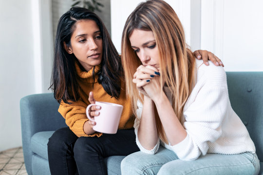 Pretty young woman supporting and comforting her sad friend while sitting on the sofa at home.