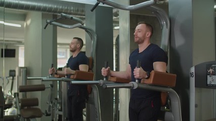 Handsome muscular built man performing hanging raises exercise on pull-up machine in health club. Male bodybuilder flexing abdominal muscles, doing straight leg raises workout on parallel bar.