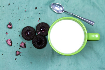 Top view of milk in green ceramic cup, metal spoon and chocolate berry cookie on green cloth background with copy space, dark tone. 