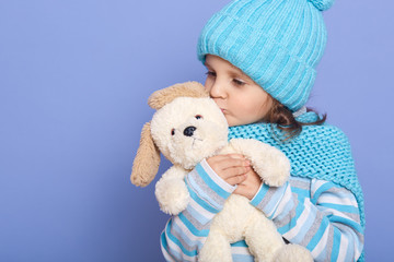 Studio shot of little charming girl wearing casually hugging and kissing her soft dog toy, posing in studio against blue wall, female child playing with her favourite toys. Childhood concept.
