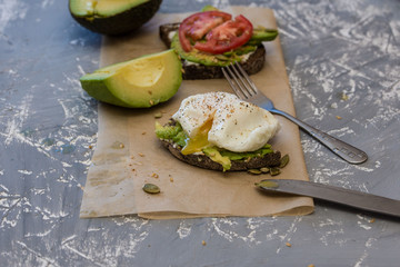 Delicious toast of avacado, black bread, flax seeds, pumpkin, juicy cucumber, tomato, poached egg on parchment with a dark background.