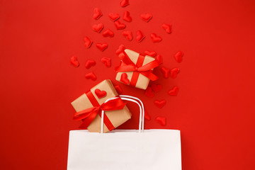 White shopping paper bag with red hearts confetti and gifts on red background. Valentine day concept. Top view, flat lay