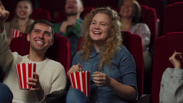 Tracking left medium shot of laughing young people enjoying watching comedy movie and eating popcorn sitting on comfortable seats in cinema