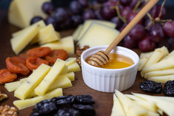 Close up photo of a board with sliced different types of cheeses with honey, grapes, nuts and dried fruits. A set of cheese plates for a party and guests on a dark background.