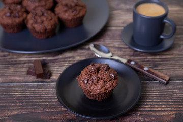Chocolate muffins on a black plate. Homemade baking. In the background is a cup of coffee and a plate with muffins. Wooden background. Selective focus, close up.