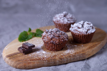 Chocolate muffins on a wooden board, strewed with icing sugar. Homemade baking. In the background is a cup of coffee and a plate with muffins. Wooden background. Selective focus, close up.