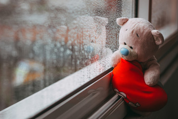 A toy pink sad bear with a red plush heart sitting on the windowsill. Autumn rainy day. Raindrops...