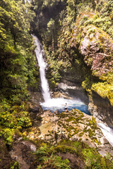 Tourist enjoying his time at beautiful Cascade creek waterfall. Man backpacker spread his arms. Pure wild lagoon in rainforest of New Zealand.