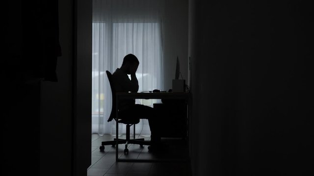 Silhouetted man in room by computer stressed, overwhelmed and fatigued.