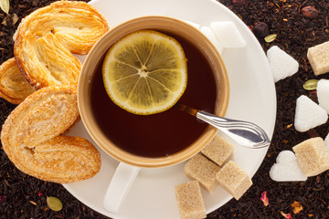 white mug with hot black tea with lemon and cookies on a white saucer on the background of black loose tea top view