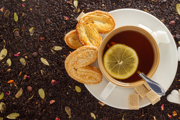 white mug with hot black tea with lemon and cookies on a white saucer on the background of black loose tea top view