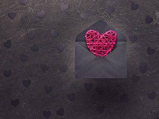 Romantic composition for Valentine's day. Red wicker heart is in a black envelope against a dark background. Decorating with paper black confetti in the form of hearts. Flat lay, copy space.