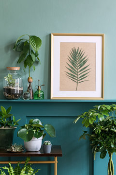 Interior design of living room with gold mock up photo frame on the green shelf with beautiful plants in different hipster and design pots. Elegant personal accessories. Home jungle. Template. 
