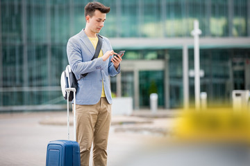 Stylish man standing at airport with suitcase and smartphone, hailing taxi.  Businessman traveling. 