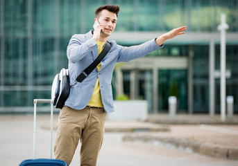 Stylish man standing at airport with suitcase and smartphone, hailing taxi.  Businessman traveling. 